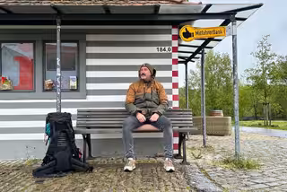 David sitzt mit Mütze und Funktionskleidung auf einer Bank vor einem kleinen Info-Häuschen. Sein Rucksack steht auf den Pflastersteinen. Neben der Bank ist ein gelbes Schild mit einem Daumen-hoch-Symbol und dem Text Mitfahrer-Bank.