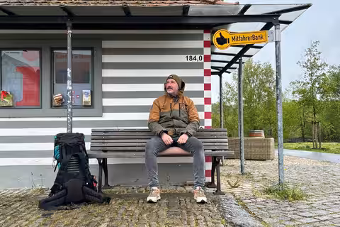 David sitzt mit Mütze und Funktionskleidung auf einer Bank vor einem kleinen Info-Häuschen. Sein Rucksack steht auf den Pflastersteinen. Neben der Bank ist ein gelbes Schild mit einem Daumen-hoch-Symbol und dem Text Mitfahrer-Bank.