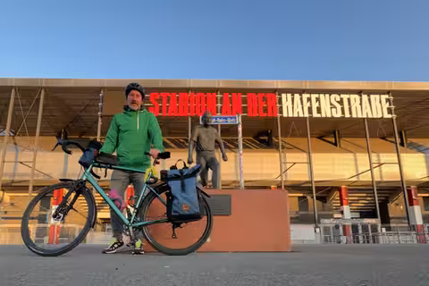 David steht mit seinem Fahrrad vor dem Stadion von Rot-Weiss Essen an der Hafenstraße. Direkt vor der Statue von Helmut Rahn. Der Himmel über dem Stadion ist wolkenlos blau.