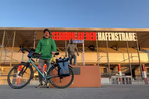 David steht mit seinem Fahrrad vor dem Stadion von Rot-Weiss Essen an der Hafenstraße. Direkt vor der Statue von Helmut Rahn. Der Himmel über dem Stadion ist wolkenlos blau.