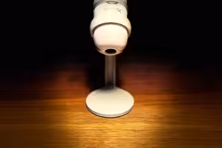 Ein weißes Mikrofon auf einem Holztisch. Nur das Mikrofon ist beleuchtet, rundherum wird alles langsam schwarz.