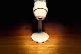 Ein weißes Mikrofon auf einem Holztisch. Nur das Mikrofon ist beleuchtet, rundherum wird alles langsam schwarz.