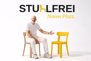 David, ganz in weiß gekleidet, sitzt auf einem Stuhl und deutet lächelnd auf einen gelben Stuhl neben ihm. Darüber steht das Logo Stuhlfrei – Nimm Platz.
