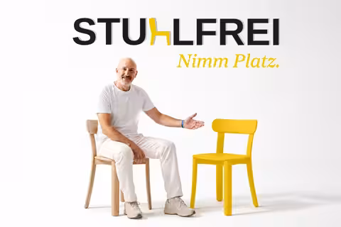 David, ganz in weiß gekleidet, sitzt auf einem Stuhl und deutet lächelnd auf einen gelben Stuhl neben ihm. Darüber steht das Logo Stuhlfrei – Nimm Platz.