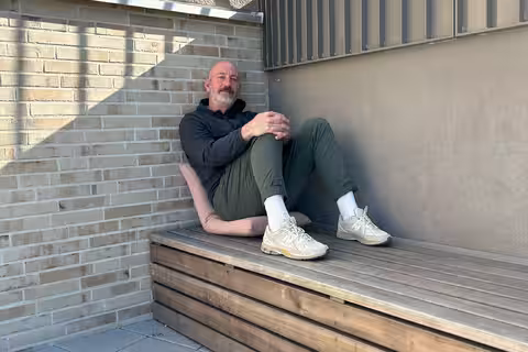 David sitzt auf seinem mitgebrachten Sitzkissen auf einer Holzbank und schaut zufrieden und entspannt in die Kamera.