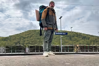 David steht mit Rucksack auf dem Rücken am Bahnhof von Gemünden (Main). Der Himmel ist etwas bewölkt.