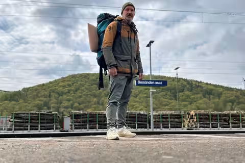 David steht mit Rucksack auf dem Rücken am Bahnhof von Gemünden (Main). Der Himmel ist etwas bewölkt.